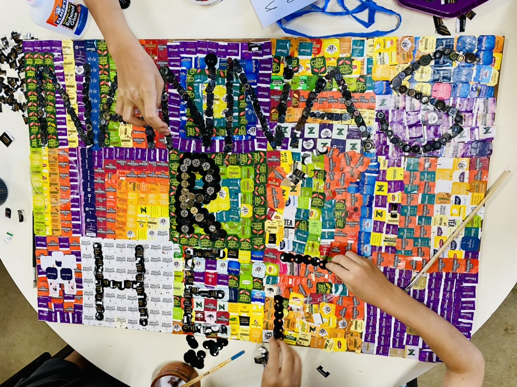 Overhead view of a table where two hands are arranging black buttons and colorful tea-tag wrappers into a mosaic spelling “Vegan Power.” Glue, paintbrushes, and scattered craft pieces surround the artwork.