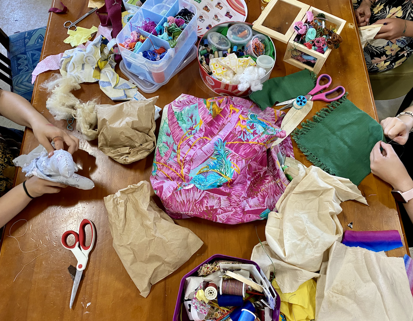 Overhead view of a wooden table covered with sewing and craft materials, including fabric scraps, thread spools, scissors, and containers of buttons and yarn. Several hands around the table are working with cloth and stuffing.