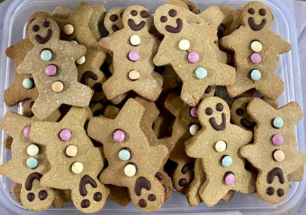 A plastic container filled with gingerbread-person cookies decorated with chocolate smiles and pastel candy buttons. The cookies are stacked and overlapping, viewed from above.