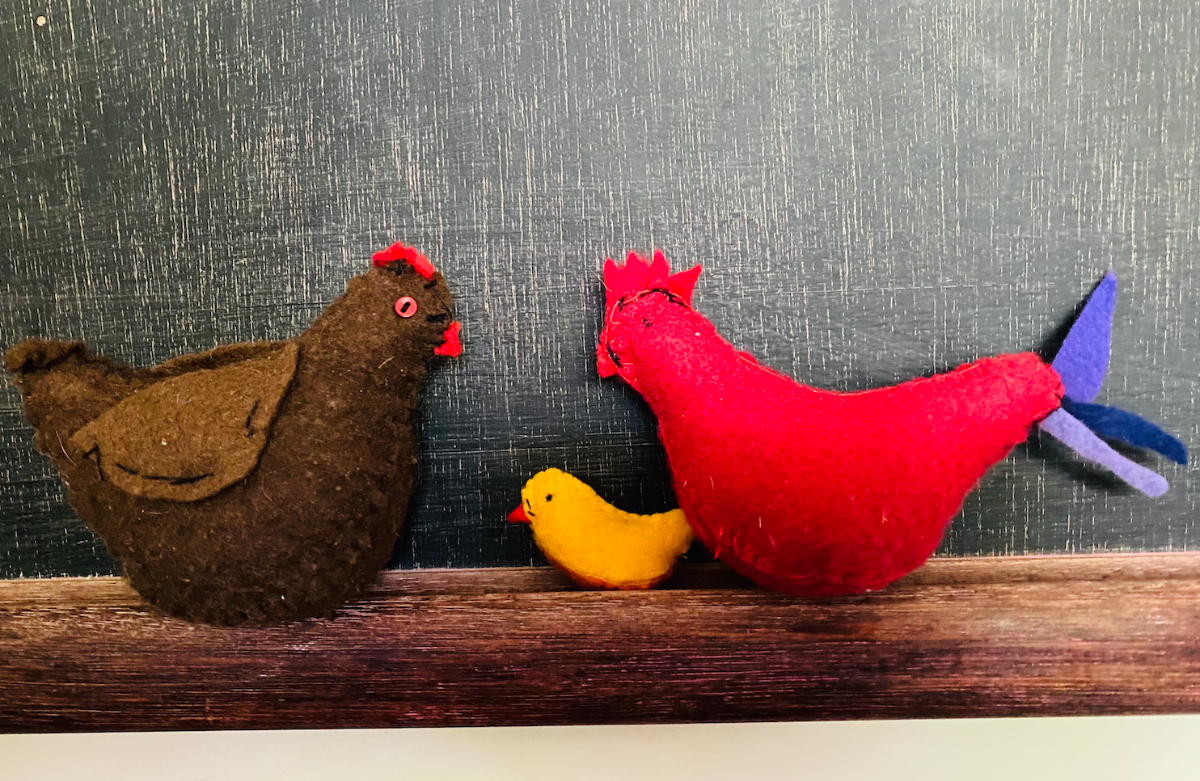 Three handmade felt birds sit on a wooden ledge against a dark textured background: a brown hen on the left, a small yellow chick in the middle, and a red hen with a purple tail on the right.