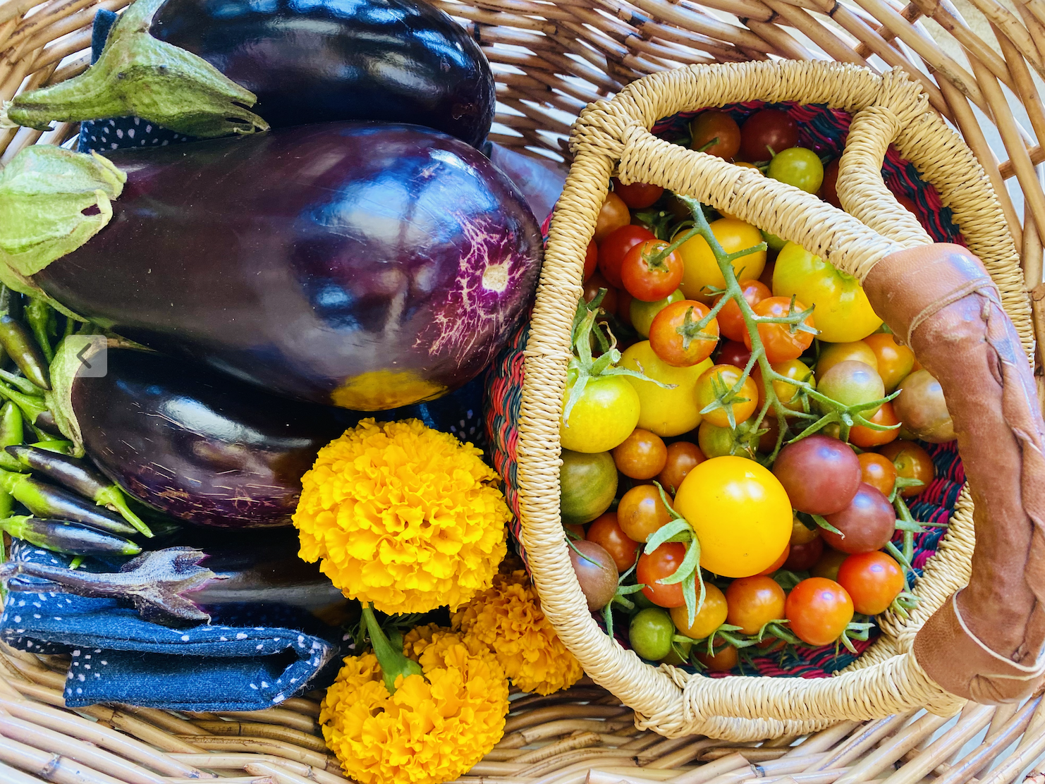 Wicker basket filled with vegetables: several large purple eggplants, a small pile of green chilies, and bright orange marigold flowers. A smaller woven tote inside holds mixed cherry tomatoes in red, yellow, and green.