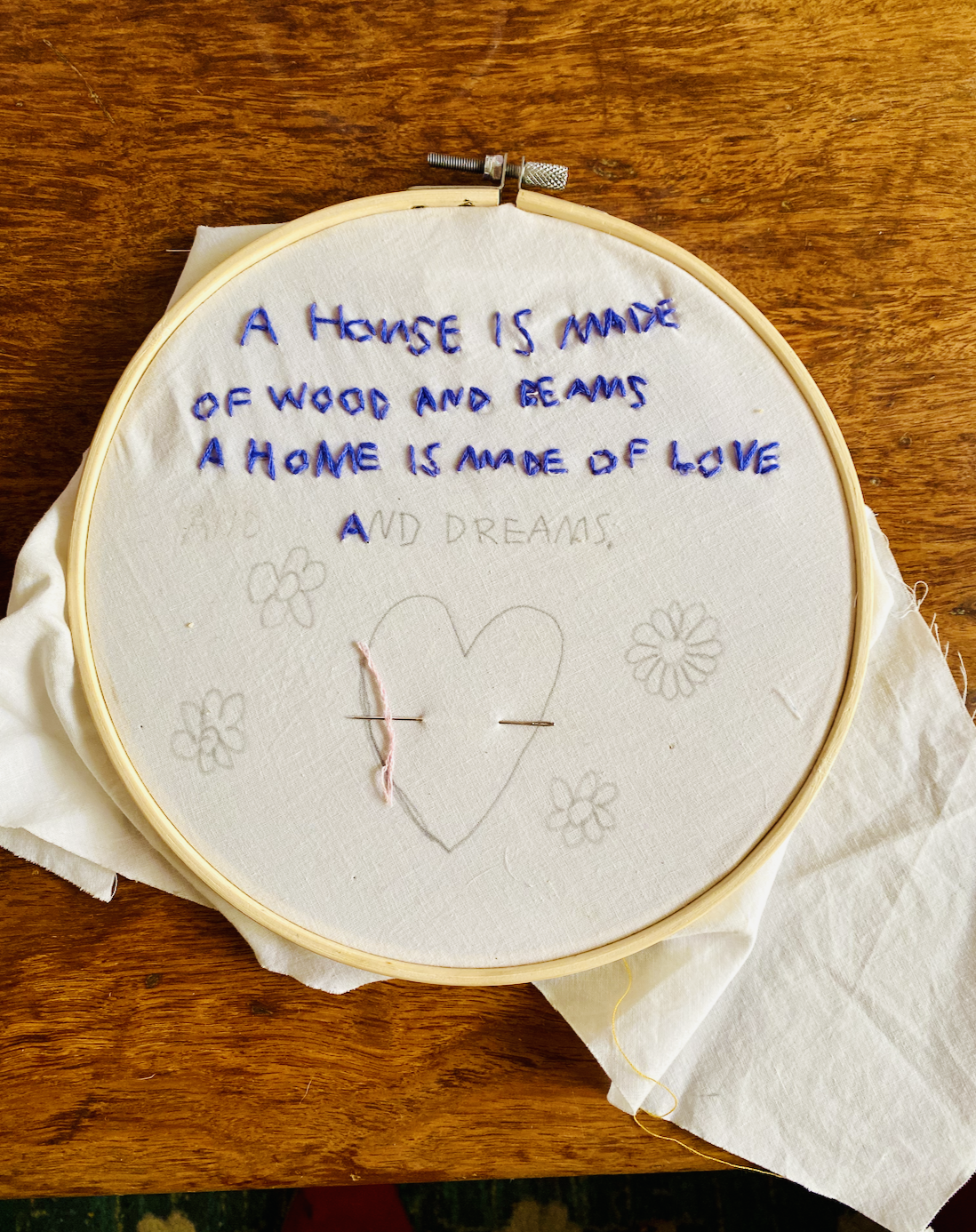 Embroidery hoop on a wooden table holding white fabric with blue stitched text reading “A house is made of wood and beams / A home is made of love,” with “and dreams” faintly penciled below. A heart and small flower shapes are sketched on the fabric, with a few stitches started in the heart.