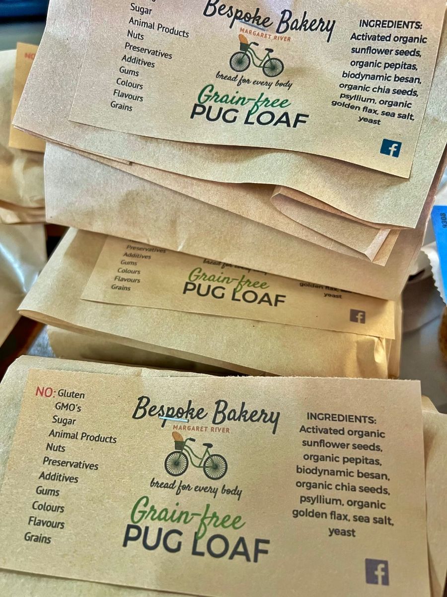 Stacked paper bread bags labeled “Bespoke Bakery” and “Grain-free Pug Loaf,” with a bicycle logo. The labels show a “NO” list (gluten, GMO’s, sugar, animal products, nuts, preservatives, additives, gums, colours, flavours, grains) and an ingredients list on the right.