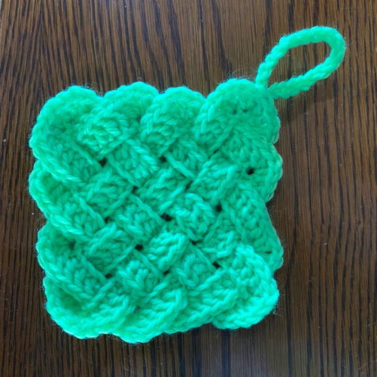 Bright green crocheted square with a textured woven pattern and a looped yarn handle in the top right. It is photographed on a dark wood-grain surface.