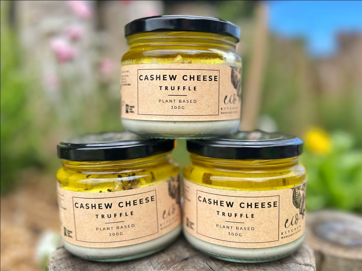Three glass jars stacked in a pyramid on a wooden surface, each with a black lid and a label reading “Cashew Cheese Truffle, Plant Based, 300g.” The jars are photographed outdoors with a blurred garden background.