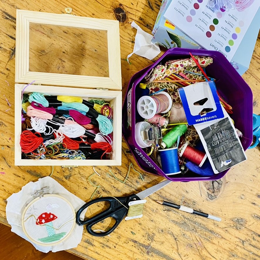 Open wooden box of colorful embroidery floss beside a purple container filled with sewing supplies like thread spools, needles, and pencils on a wooden table. A small embroidery hoop with a stitched red-and-white mushroom and a pair of black scissors lie in the foreground.