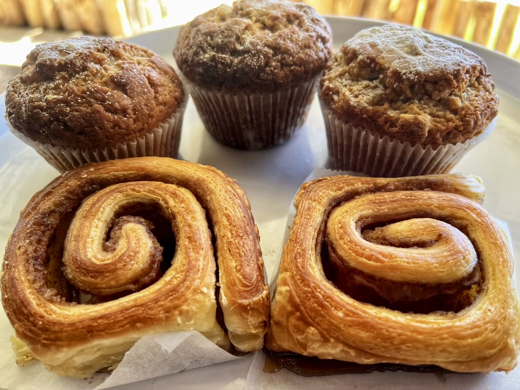Close-up of a white plate holding three muffins in paper liners and two large spiral cinnamon rolls. Warm light and a blurred wooden background are visible behind the pastries.