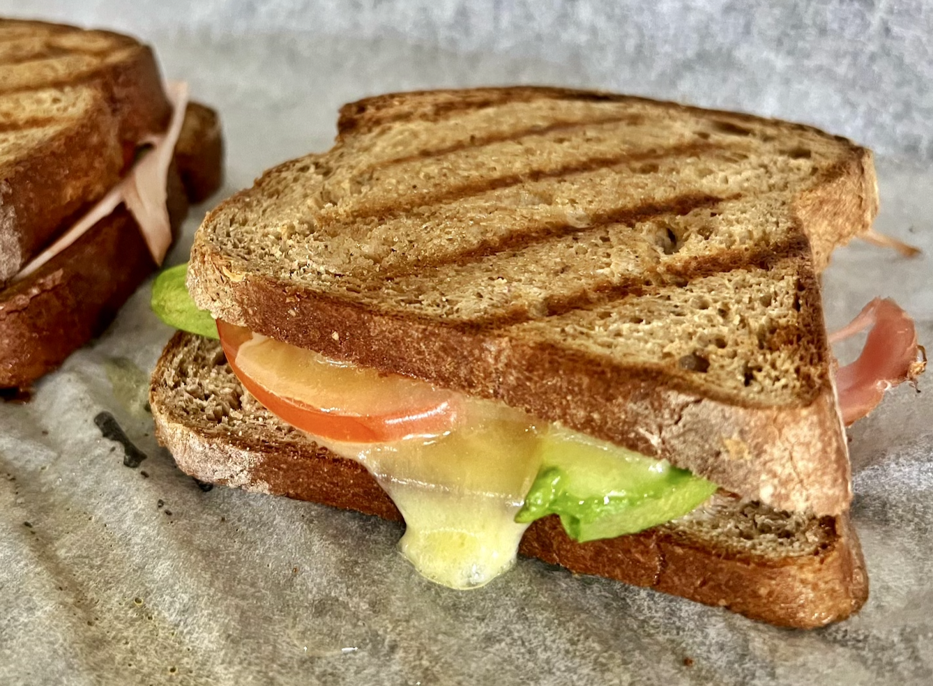 Close-up of a toasted sandwich on parchment paper with melted cheese dripping out, plus slices of tomato and avocado inside. Another sandwich is partially visible in the background.