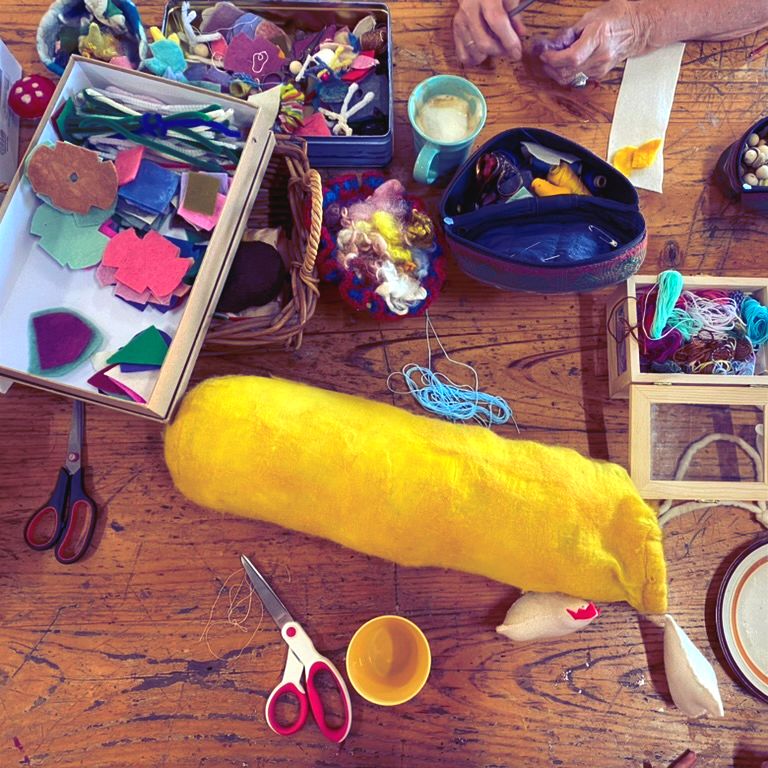 Overhead view of a wooden table covered with craft supplies, including felt cutouts, scissors, thread, a mug, and small trays and boxes. A long yellow felted shape with a white end lies across the center while hands work near the top right.