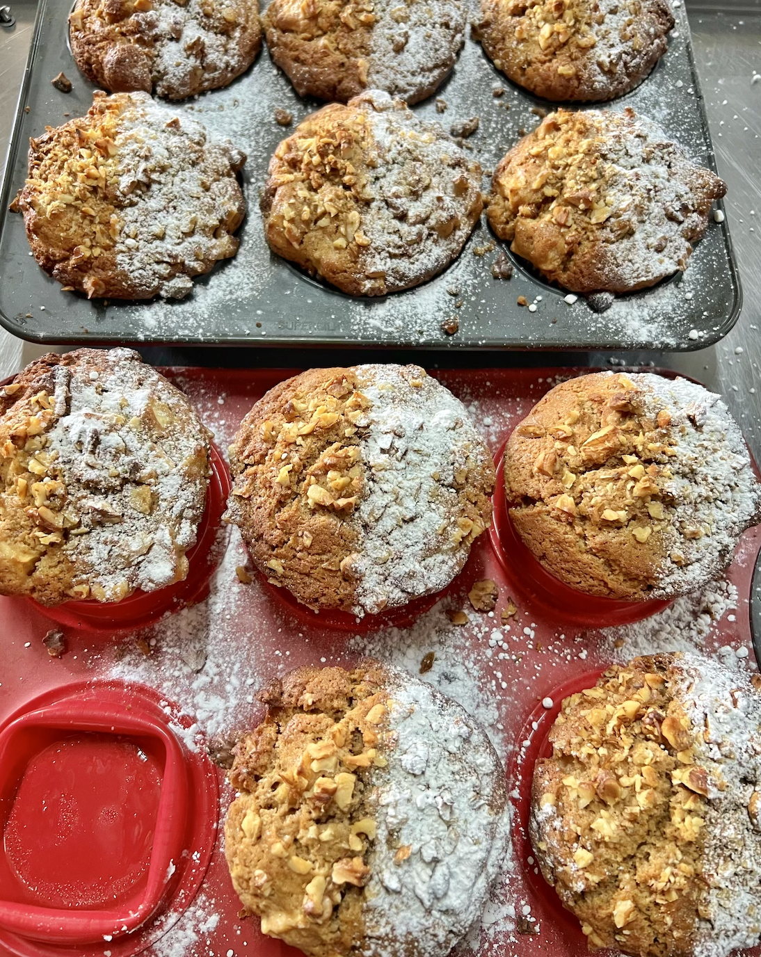 Freshly baked muffins topped with chopped nuts and dusted with powdered sugar sit in a metal muffin tray and red silicone baking cups. Powdered sugar is scattered across the trays and work surface.