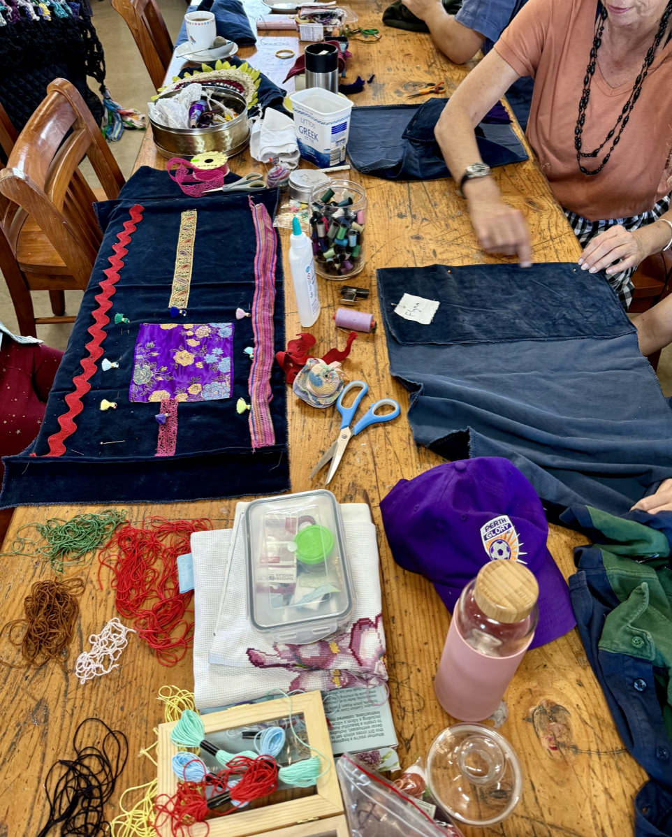 Craft supplies cover a wooden table, including denim fabric pieces, scissors, threads, ribbons, glue, and a jar of markers. Several people sit around the table working with fabric, with a purple cap and a pink water bottle near the front.