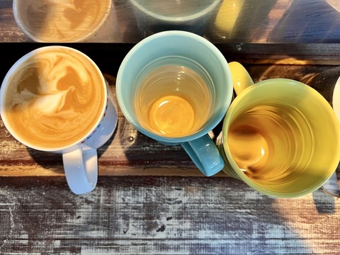 Overhead view of three mugs on a weathered wooden table: a light blue mug with coffee and foam, a teal mug with a small amount of amber liquid, and a green mug filled with golden tea. Sunlight casts warm highlights and shadows across the cups and tabletop.