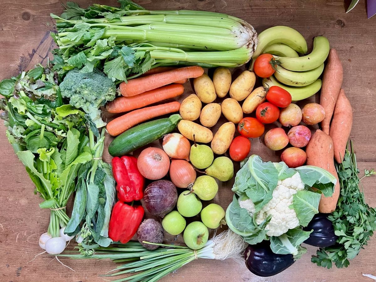 Assorted produce arranged on a wooden table, including celery, carrots, broccoli, potatoes, tomatoes, bananas, peppers, onions, pears, apples, cauliflower, eggplants, and mixed greens. Spring onions, herbs, and a few root vegetables are grouped along the edges.