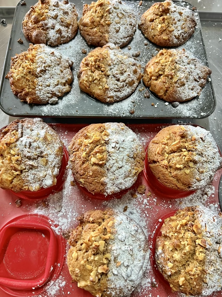 Tray of large golden-brown muffins topped with chopped nuts and dusted with powdered sugar. Several muffins sit on a red surface below, with powdered sugar scattered around.