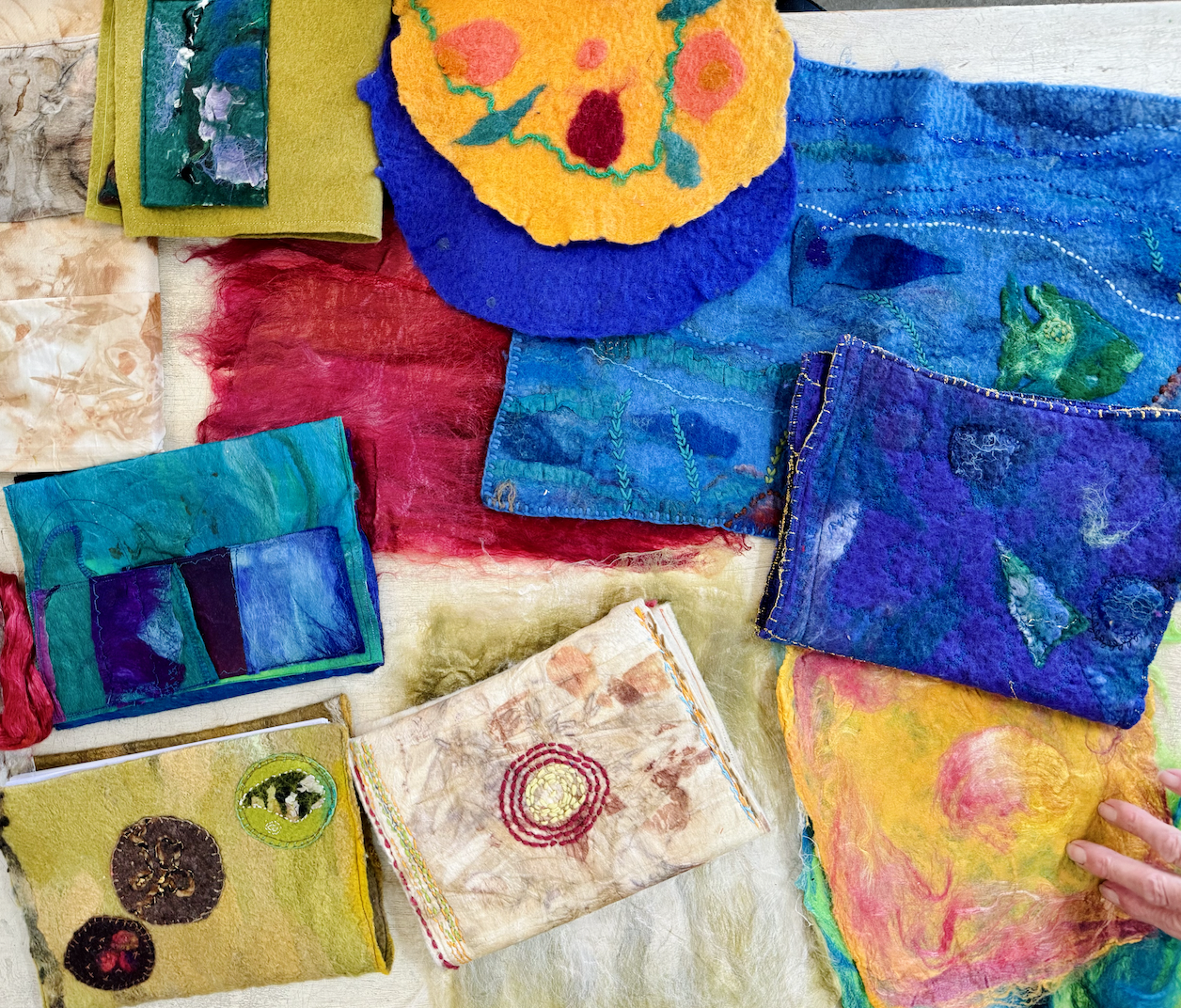 Colorful handmade felt and fabric pieces—stitched pouches, panels, and a round flower-like felt piece—layered on a light surface. A hand at the lower right touches a yellow-and-pink felt sheet.