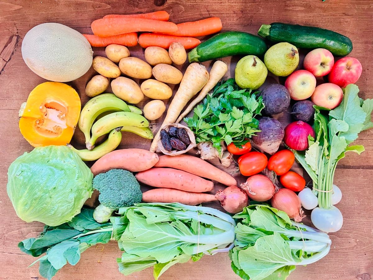 Assorted produce arranged on a wooden table, including carrots, potatoes, zucchini, pears, apples, bananas, tomatoes, onions, beets, and leafy greens. Also shown are a melon, a halved squash, broccoli, parsnips, ginger, dates, and white turnips.