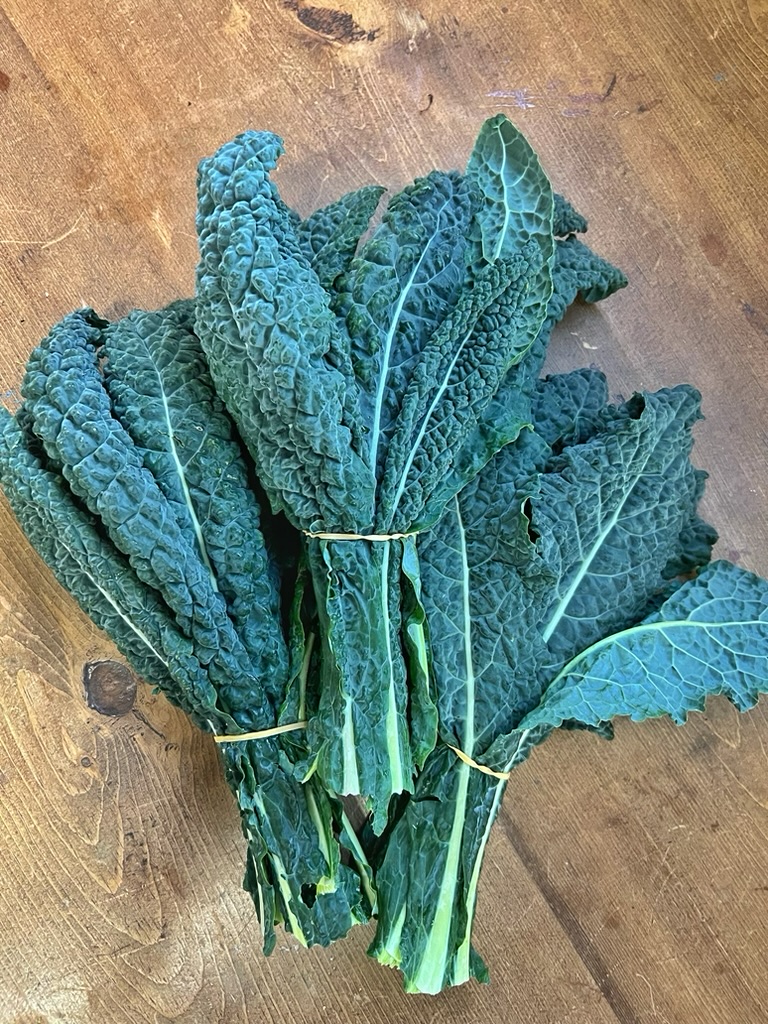 Bundle of dark green, crinkled kale leaves tied with a rubber band on a wooden tabletop. The stems and leaf veins are visible in a top-down view.