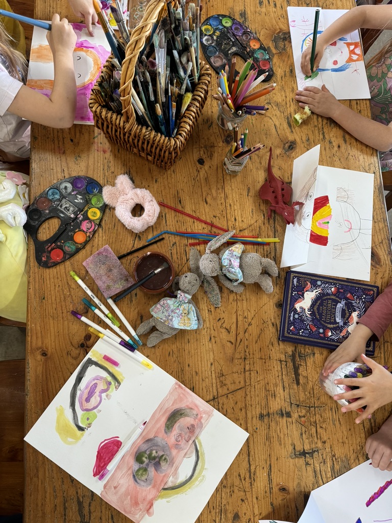 Overhead view of children’s hands drawing and painting at a wooden table covered with sketchbooks, watercolor palettes, markers, and brushes. A woven basket of paintbrushes, jars of colored pencils, two stuffed bunnies, and a small red toy dinosaur sit among the art supplies.