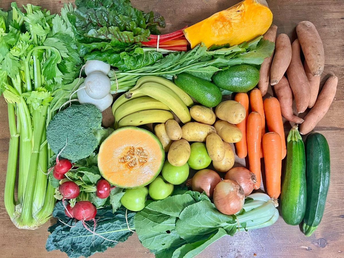 Assorted produce on a wooden table, including celery, leafy greens, broccoli, radishes, turnips, a halved melon, bananas, green apples, potatoes, onions, carrots, avocados, and cucumbers. A wedge of squash and several sweet potatoes are arranged along the top right.
