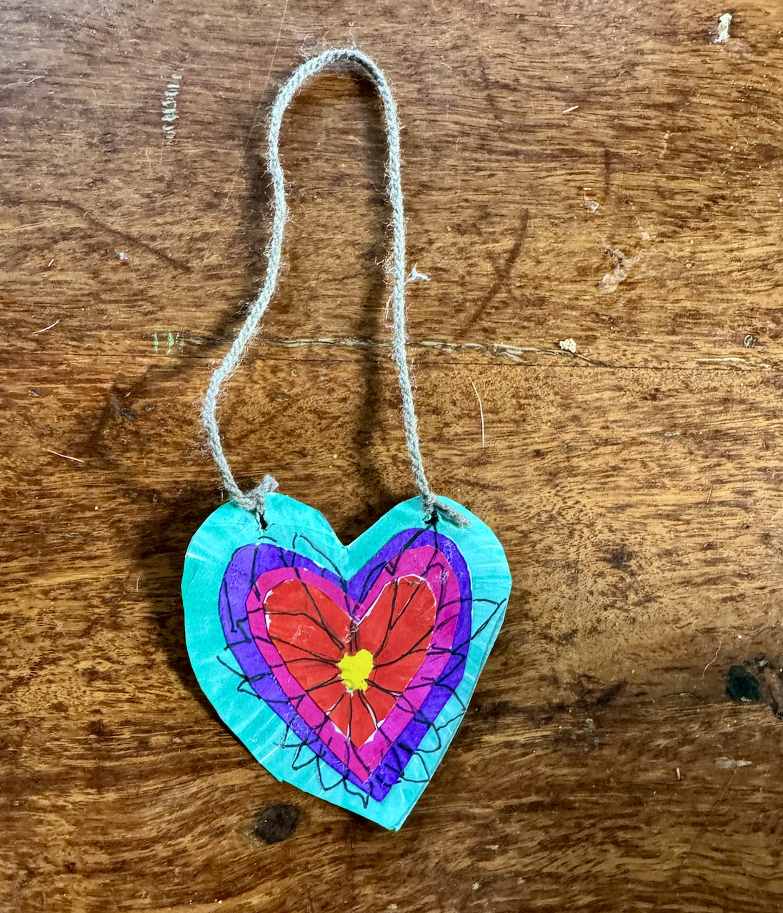 Handmade heart-shaped ornament painted turquoise with layered purple, pink, and red heart designs, tied with a loop of twine. It lies on a worn wooden tabletop.