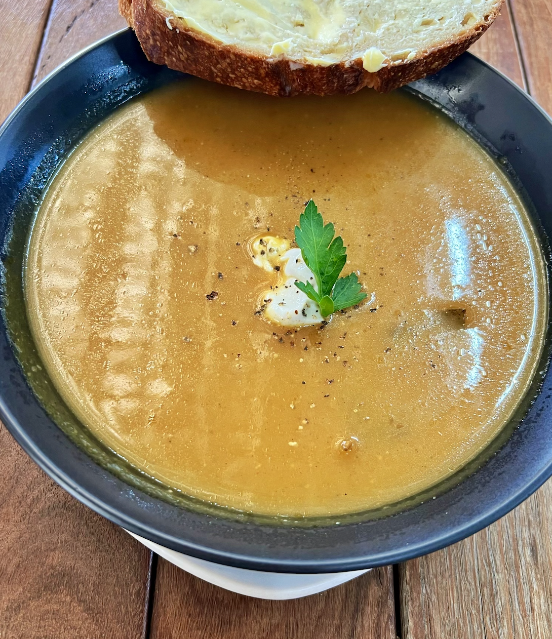 A black bowl filled with golden-brown soup sits on a wooden table, garnished with a parsley leaf and a few drops of cream and pepper. A buttered slice of bread rests on the rim of the bowl.