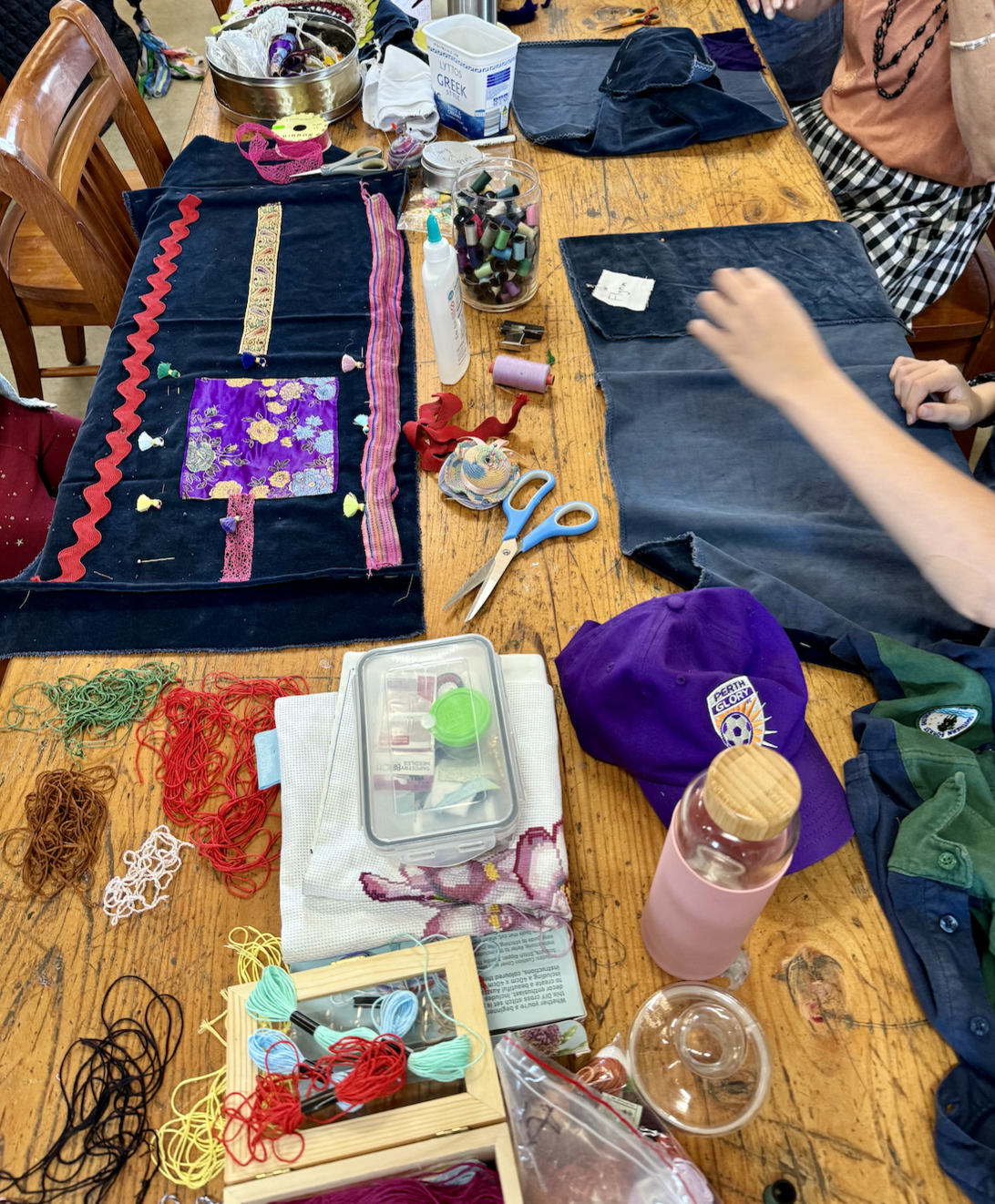 Wooden table covered with sewing and craft supplies, including fabric pieces, ribbons, thread, scissors, pins, and a jar of spools. A hand reaches toward a dark fabric panel while another decorated fabric piece lies beside it.
