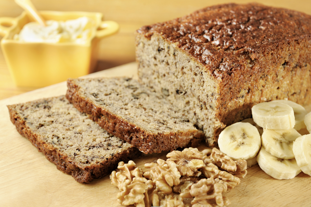 A loaf of banana bread on a wooden cutting board with two slices cut in front. Banana slices and walnut pieces sit beside the loaf, with a yellow dish in the blurred background.