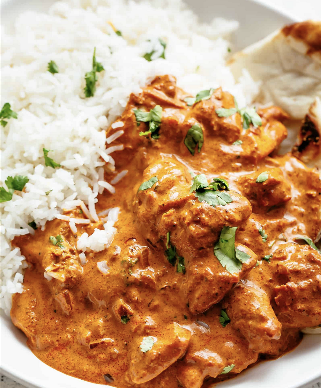 Close-up of a white plate with fluffy white rice alongside chunks of meat in a creamy orange-red sauce, topped with scattered green herb leaves. A piece of flatbread is visible at the edge of the plate.