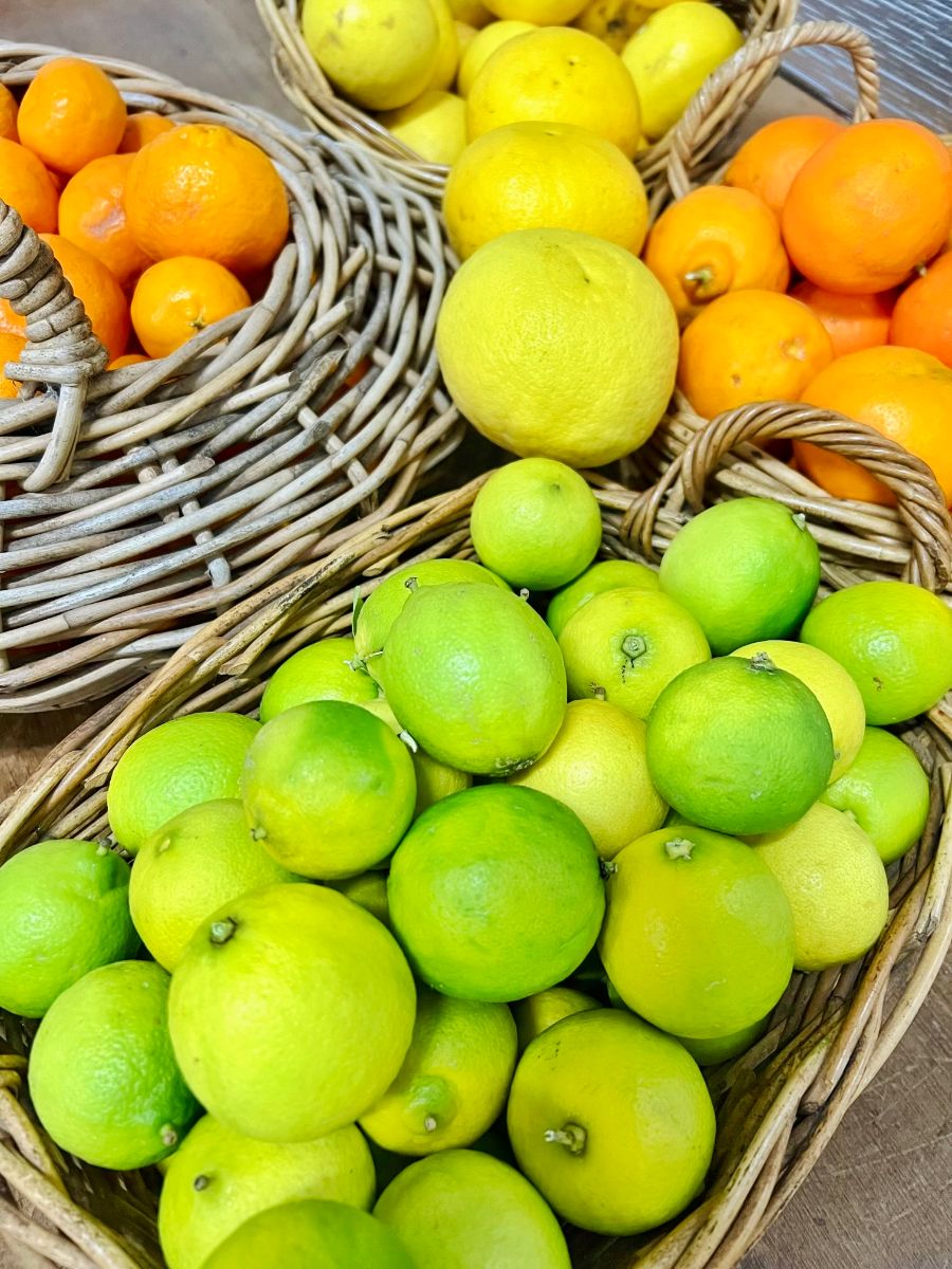 Wicker baskets filled with citrus fruit, including bright green limes in the foreground. Yellow lemons and orange-colored citrus sit in baskets behind on a wooden surface.