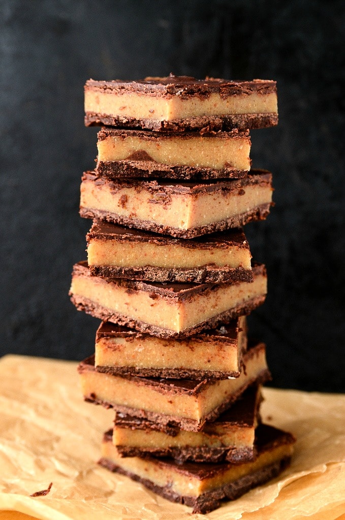 A tall stack of rectangular caramel slice bars with chocolate tops and bases sits on parchment paper. The background is dark and out of focus.