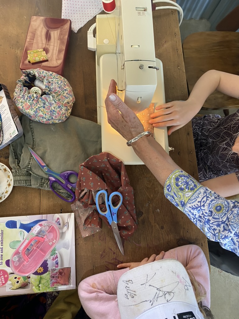 Overhead view of a child and an adult using a sewing machine at a wooden table. Scissors, fabric pieces, a pincushion with sewing supplies, and a craft kit are spread around the machine.