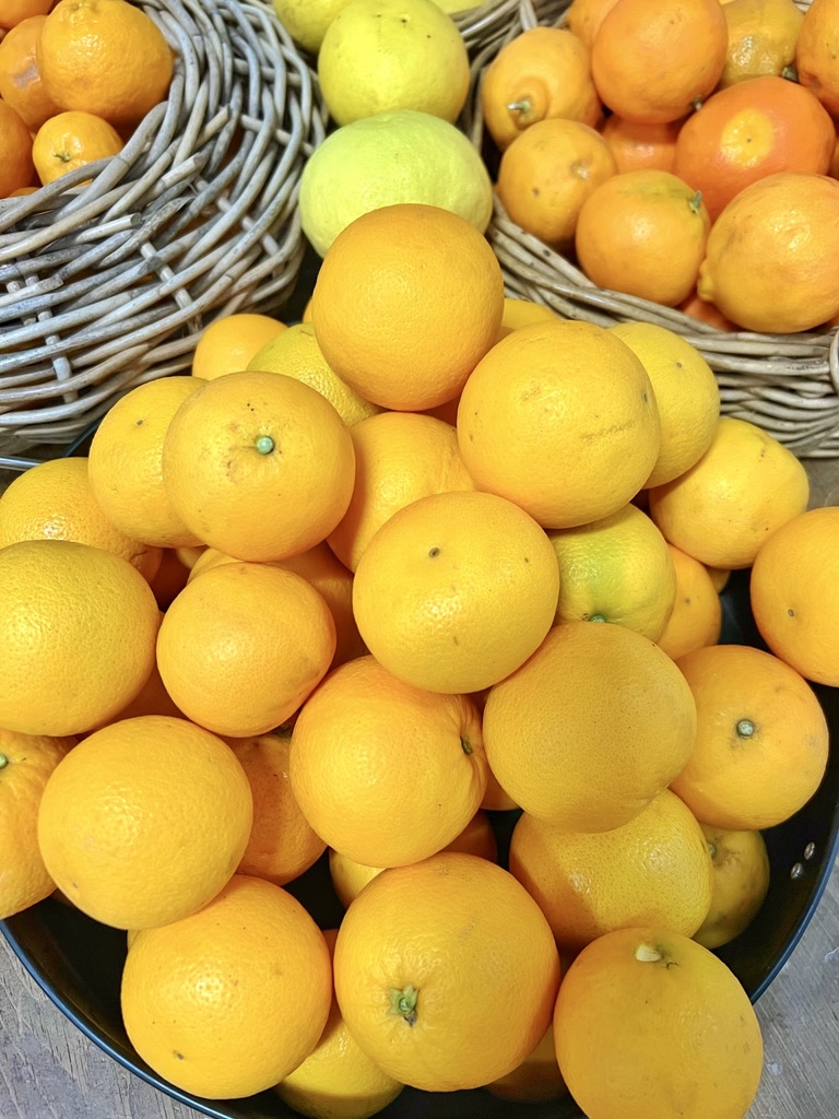 Close-up of wicker baskets and a metal tub filled with citrus fruit, mostly oranges with a few yellow and green fruits mixed in. The round fruits are piled high, with basket rims visible around the edges.