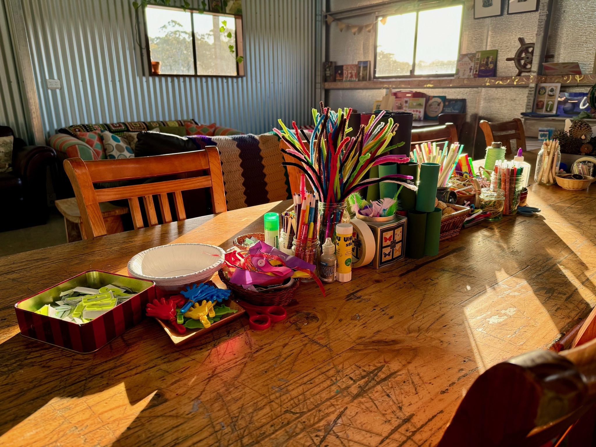 Sunlit wooden table set up with kids’ craft supplies, including jars of colorful pipe cleaners, pencils and markers, scissors, glue, paper plates, and small baskets. In the background are wooden chairs, a patterned couch with blankets, and windows and shelves inside a corrugated-metal room.