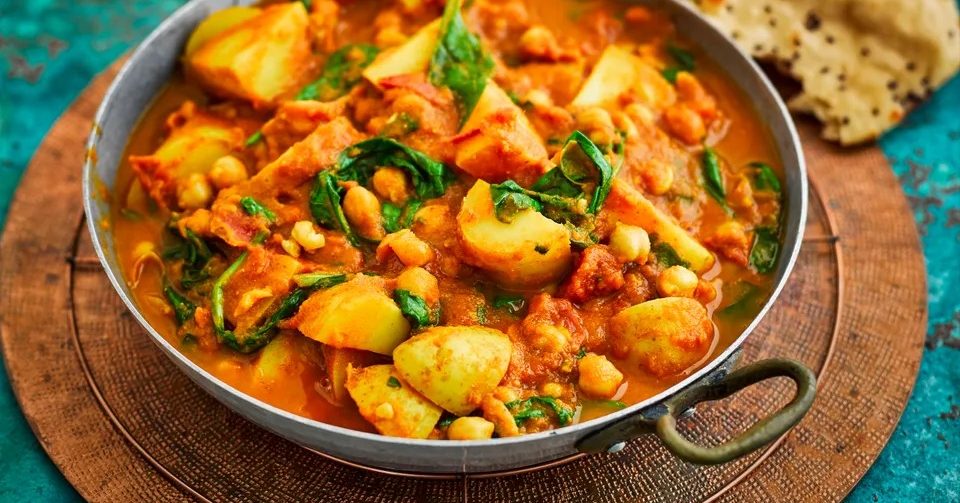 Close-up of a metal pan filled with orange-red stew containing potato chunks, chickpeas, and leafy greens. The pan sits on a round wooden board on a teal surface, with a piece of flatbread in the corner.
