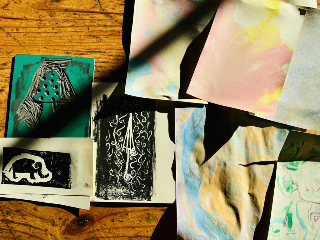 Assorted prints and marbled papers are spread on a wooden table. The pieces include black-and-white block-print designs (one resembling an elephant) and colorful pastel sheets with swirling patterns.