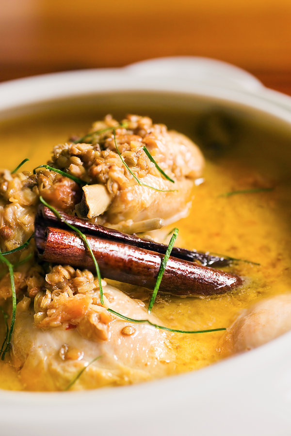 Close-up of a white bowl filled with golden broth, topped with dumpling-like pieces and grains. A cinnamon stick and thin green herb strands rest on top.