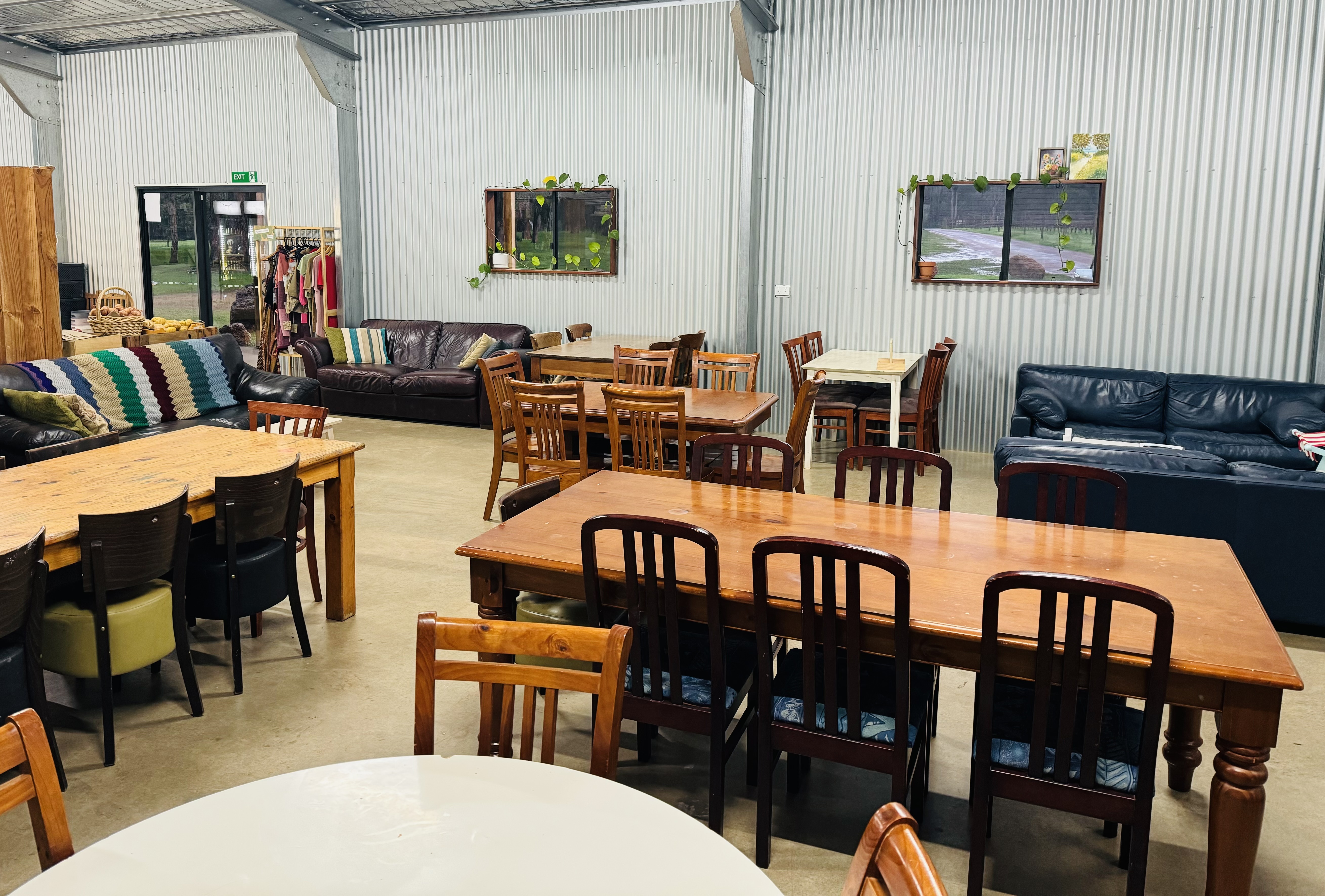 Interior of a corrugated-metal café seating area with several wooden tables and mismatched chairs, plus leather couches along the walls. Two windows with trailing plants and a glass door are visible in the background.