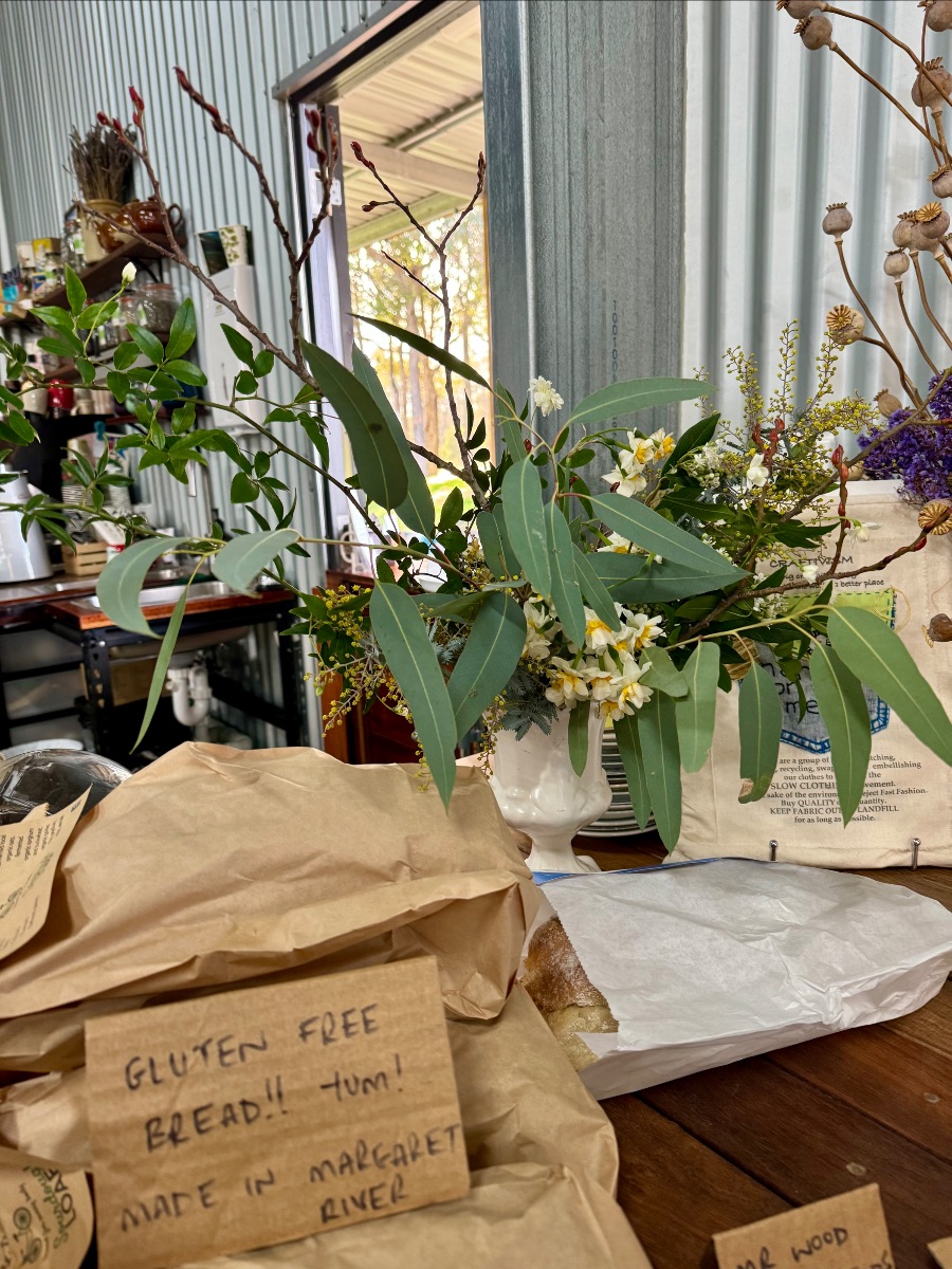 Paper-wrapped baked goods sit on a wooden table, including a handwritten sign reading “Gluten free bread!! Yum! Made in Margaret River.” Behind them, a large leafy bouquet with white flowers in a white vase stands in front of a corrugated metal wall and open doorway.