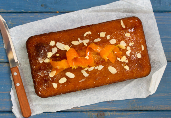 Rectangular loaf cake on a sheet of parchment paper, topped with orange peel pieces and sliced almonds. A serrated knife with a wooden handle lies to the left on a blue wooden surface.
