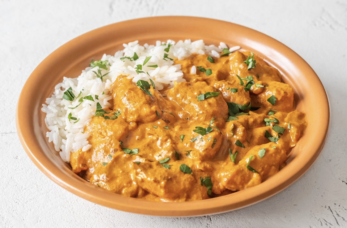 A shallow orange bowl filled with white rice and chunks of food in a creamy orange sauce, topped with chopped green herbs. The bowl sits on a light gray surface.