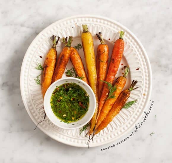 Overhead view of a white plate holding roasted carrots with short green tops around a small bowl of green herb sauce. A few carrot fronds are scattered on the plate and marble surface.