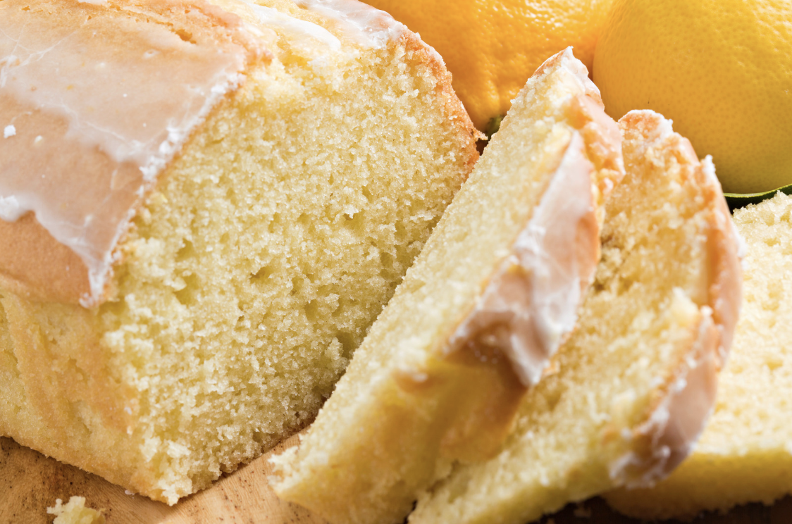 Close-up of a glazed loaf cake with several slices cut and laid in front on a wooden board. Whole lemons sit in the background.