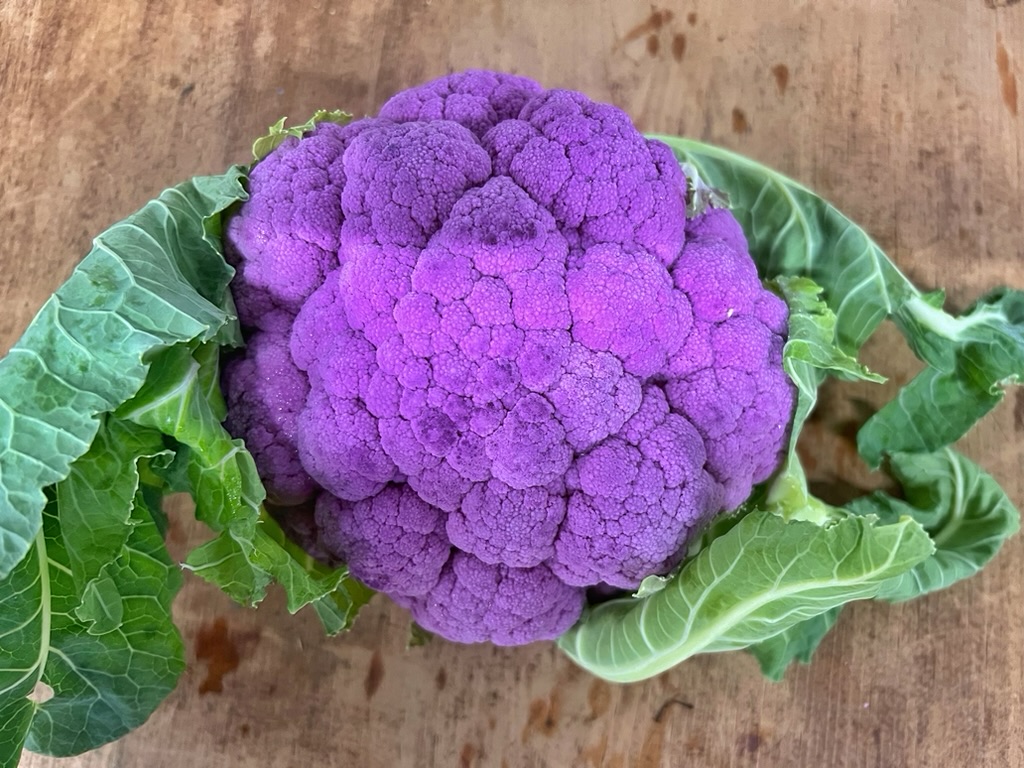 Close-up of a bright purple cauliflower head with green leaves around it. The cauliflower sits on a wooden surface.