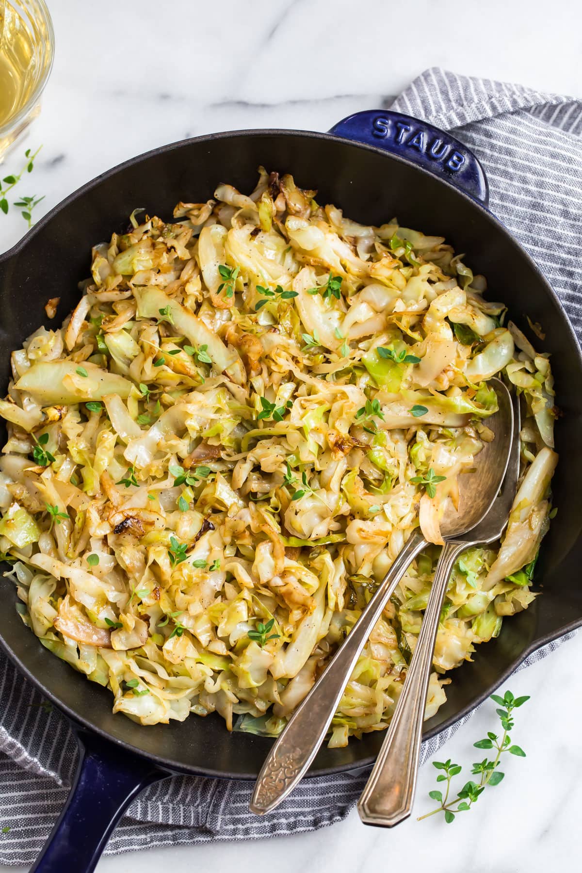 Overhead view of a black skillet filled with sautéed chopped cabbage, garnished with small green herb leaves. Two metal serving spoons rest in the pan on a striped dish towel, with a light countertop and a glass of pale liquid partially visible.