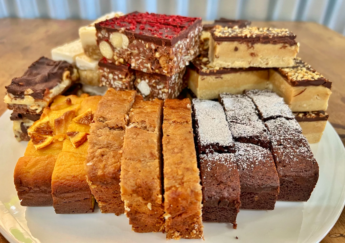 Assorted dessert bars and slices stacked on a white plate, including chocolate-topped squares with nuts, caramel-colored slices, orange-topped cake pieces, and powdered-sugar brownies. The plate sits on a wooden table with a softly blurred background.