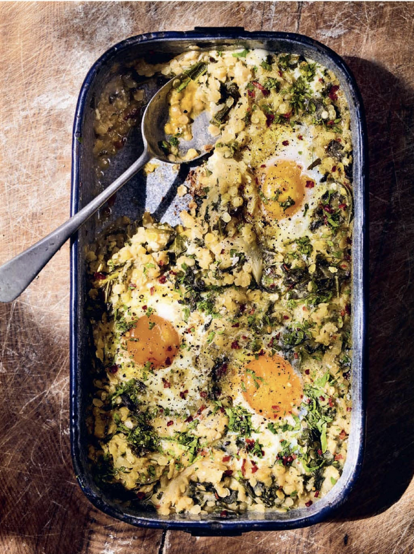 Overhead view of a rectangular baking dish filled with a lentil-and-greens mixture topped with three baked eggs and sprinkled with chopped herbs and pepper flakes. A metal serving spoon rests in the dish on a wooden surface.