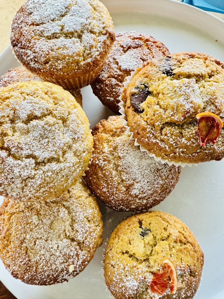 Close-up of six baked muffins on a light plate, dusted with powdered sugar. One muffin shows blueberry pieces and another has a slice of dried citrus on top.