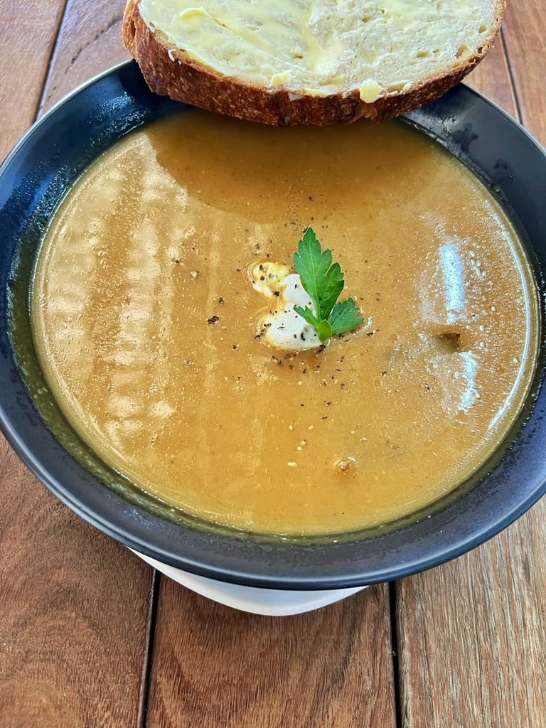 Bowl of smooth brown soup topped with a small dollop of cream, black pepper, and a parsley leaf. A slice of buttered bread rests on the rim of the bowl on a wooden table.