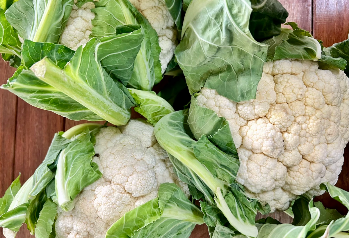 Close-up of several fresh cauliflower heads with green leaves still attached, arranged on a wooden surface. The pale florets and leafy stems fill most of the frame.