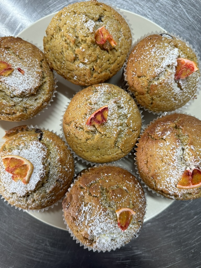 Seven golden-brown muffins in paper liners sit on a white plate on a metal surface. They’re dusted with powdered sugar and topped with small slices of red-orange citrus.